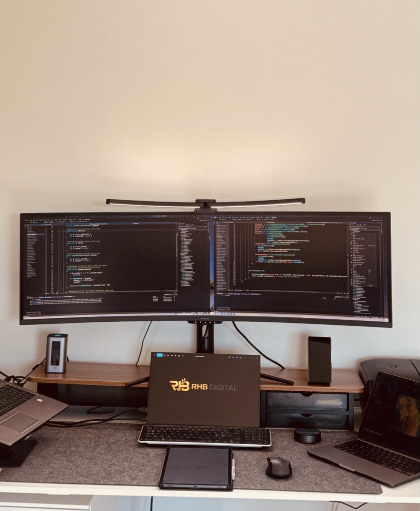 Workspace with a curved dual-monitor computer setup showing code, on a standing monitor arm above a keyboard and laptop.