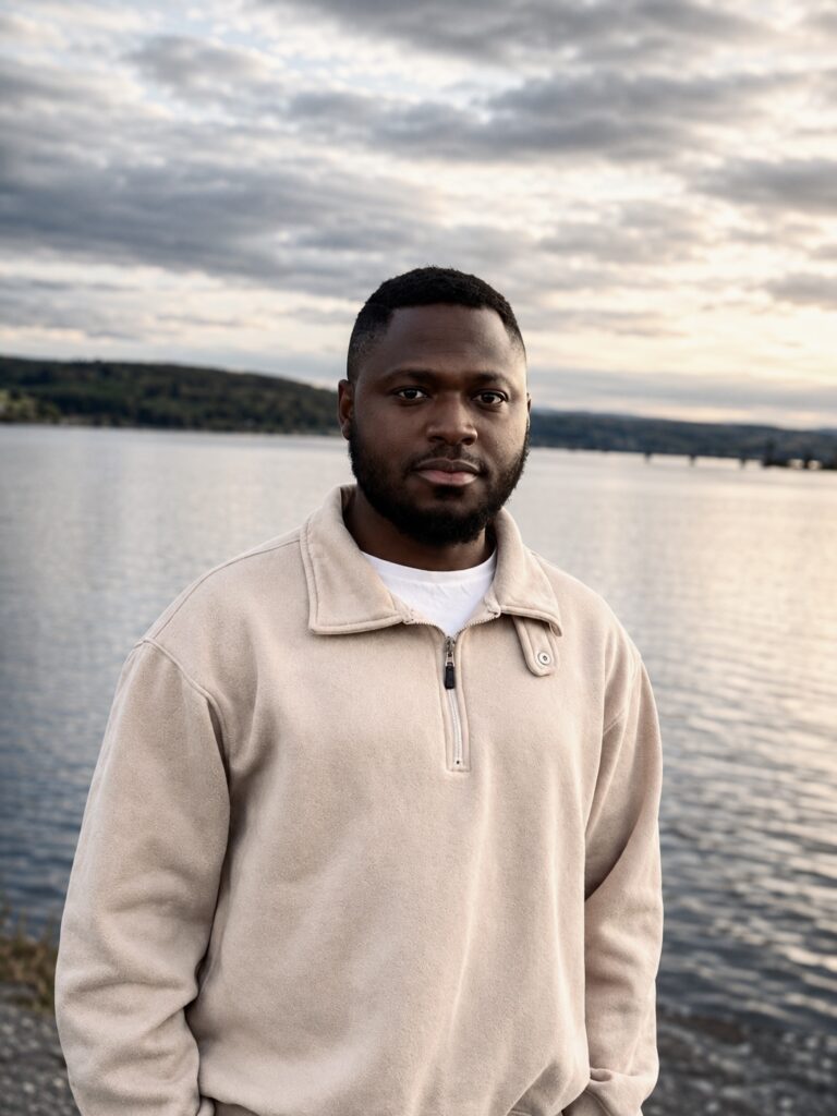 Adult man standing near a lake with a calm water backdrop and cloudy sky, wearing a beige zip-up fleece jacket.